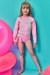 Menina sorridente vestindo um biquíni e blusa em malha cirrê colorida, com proteção UV 50+, em fundo azul e objetos infláveis rosa ao redor.