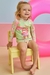 Criança sorridente sentada em uma cadeira, vestindo um conjunto de blusa e calcinha em malha com proteção UV 50+, decorado com estampas coloridas.