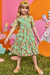 Menina sorrindo vestindo um vestido infantil Kukiê em tule, com estampas florais em tons de verde e laranja, em um cenário colorido.