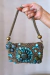 Bolsa Tesouro Turquesa decorada com pedras em tons de azul e verde, segurada por uma mão.