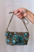 Bolsa Tesouro Turquesa com detalhes em pedras e contas em tons de turquesa, segurada por uma mão.