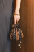 Bolsa bucket listrada em tons de marrom e preto, com detalhes em lantejoulas e alça de cordão, sendo segurada por uma mão.