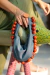 Uma mulher segurando uma bolsa de tecido azul com contas vermelhas, enquanto está sentada em um objeto decorativo de forma de animal.