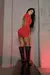 Mulher com vestido curto vermelho de gola alta e costas abertas, usando botas pretas de cano alto, posando em ambiente com parede cinza