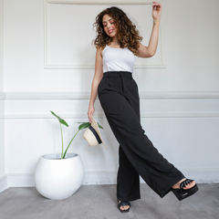 Pantalón sastrero negro de mujer, con blusa blanca, posando con un sombrero y una planta decorativa.