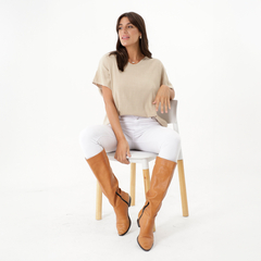 Mujer sentada en una silla, luciendo una blusa beige y pantalones blancos, con botas largas color marrón.