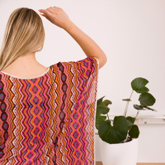 Vista trasera de una mujer con blusa de estampado colorido y ondulado, con cabello largo. En el fondo, una planta en maceta.