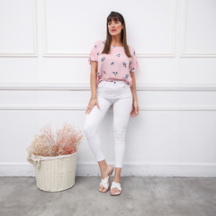 Modelo usando una blusa rosa a rayas con flores, combinada con pantalones blancos y sandalias. Fondo minimalista.
