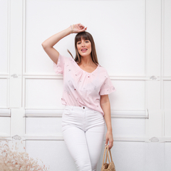 Mujer posando con una blusa rosa con detalles bordados, pantalones blancos y una bolsa de mano, en un fondo blanco.