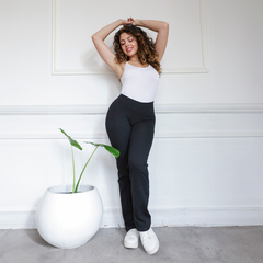 Modelo usando calza básica recta negra, combinada con una camiseta blanca, sonriendo y posando en un ambiente luminoso con una planta decorativa.