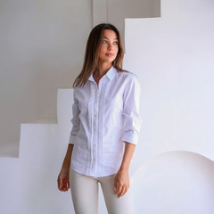 Modelo usando una camisa blanca Cleo de manga larga, con detalles en el frente, posando frente a una pared blanca.