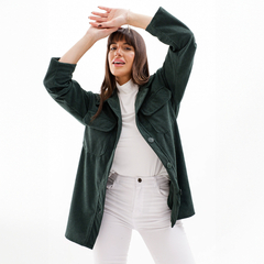 Mujer con un camisaco verde oscuro, sonriendo y posando con los brazos levantados, usando una camiseta blanca y pantalones blancos.