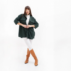 Mujer posando con un camisaco verde en un fondo blanco, usando pantalones blancos y botas marrones.