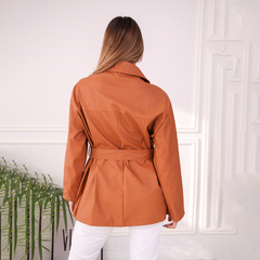 Vista posterior de un camisaco trench color marrón, con cinturón, sobre una modelo. Fondo con detalles arquitectónicos y planta decorativa.