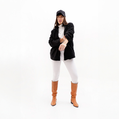 Modelo usando una campera negra con pantalones blancos y botas marrones, posando con una mano en la muñeca.