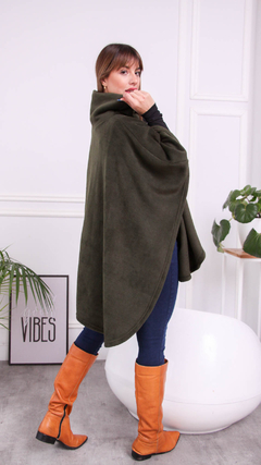 Mujer usando una capa Marano de color verde oliva, combinada con pantalones ajustados y botas altas de cuero naranja, posando en un entorno moderno.