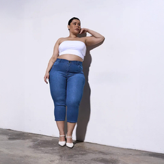 Mujer de pie con top blanco y jeans azul, posando contra una pared blanca.