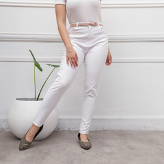 Una mujer de pie usando jeans blancos con un cinturón rosa, en un ambiente moderno con una planta decorativa.
