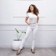 Mujer sonriente usando jeans blancos y camiseta blanca, posando en un entorno moderno con una planta en maceta.
