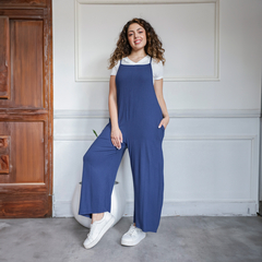 Modelo posando con un mono azul marino, de tirantes y pantalones anchos, combinado con una camiseta blanca y zapatillas. Fondo con detalles arquitectónicos.