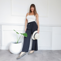 Mujer posando con pantalones Palazzo Nina de color negro, camiseta blanca y sombrero, junto a una planta en maceta.