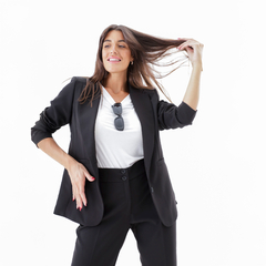 Mujer sonriente usando un blazer negro, posando con una mano en el cabello y gafas de sol en la cabeza.