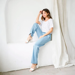 Mujer sentada en una pared, vistiendo un pantalón Lorena de color celeste y una blusa blanca, con zapatos de tacón blancos.