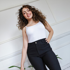 Mujer con cabello rizado usando un pantalón recto negro y una camiseta blanca, sonriente en un entorno luminoso.
