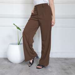 Pantalón sastrero de color marrón, con corte elegante, combinado con una blusa blanca y sandalias negras, de pie junto a una planta decorativa.