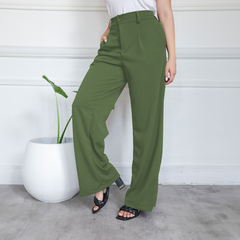 Pantalón sastrero verde, de corte elegante, usado por una mujer que posa junto a una planta decorativa.