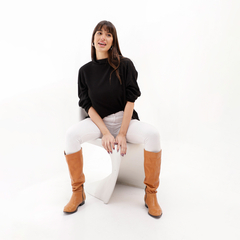 Modelo usando una polera negra de lanilla, sentada en una silla blanca, con pantalones blancos y botas marrones.