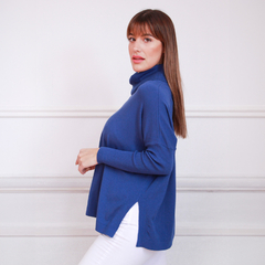 Modelo luciendo un polerón azul con cuello alto y detalles en los laterales, combinado con pantalones blancos.