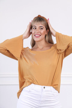 Modelo luciendo una remera larga color mostaza con detalles brillantes en el pecho, combinada con pantalones blancos y un accesorio en el cabello.
