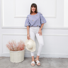Remera Kaia de color azul con bordados, combinada con pantalones blancos y sandalias. La modelo sostiene un sombrero blanco y está de pie junto a una canasta con flores secas.