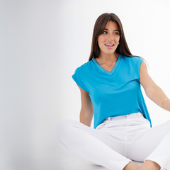 Modelo usando una blusa azul con detalles en el escote, sentada sobre el suelo con pantalones blancos.