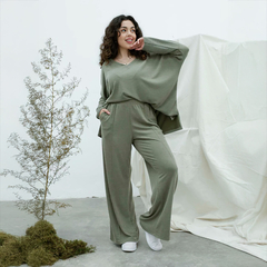 Mujer sonriente usando un remerón color verde oliva, combinado con pantalones anchos, de pie junto a una planta pequeña en un fondo neutro.