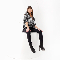 Mujer sentada sobre una superficie blanca, vistiendo una blusa negra con estampado blanco y pantalones negros, con botas de tacón.
