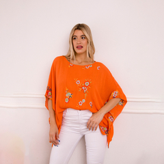Remerón naranja con bordados florales, modelo femenino que viste un pantalón blanco, fondo neutro.