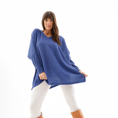 Mujer posando con un remerón azul sobre fondo blanco, usando pantalones blancos y botas marrones.