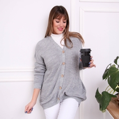 Mujer sonriente usando un saco inglés gris con botones, sosteniendo un vaso de café, de pie junto a una planta.