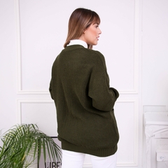 Mujer de espaldas usando un saco inglés de color verde oliva, con un fondo minimalista y una planta decorativa al lado.