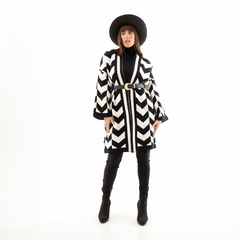 Modelo luciendo un saco de diseño en zigzag blanco y negro con cinturón, junto a un sombrero negro.