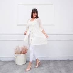 Modelo usando un spolverino elegante de color crema con detalles bordados, combinado con pantalones blancos y sandalias doradas.