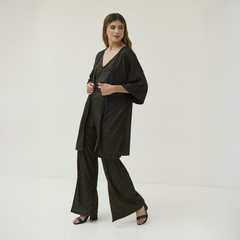 Modelo usando un spolverino negro con brillo, combinado con pantalones y sandalias, en un fondo blanco.