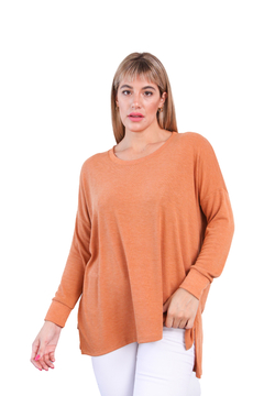 Sweater largo de color naranja, modelo Greta, con manga larga y corte asimétrico, lucido por una mujer. Se combina con pantalones blancos.