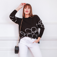 Modelo luciendo un suéter negro con diseños florales en blanco, combinado con pantalones blancos y un bolso negro.