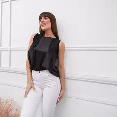 Musculosa negra con detalles de volantes y diseño con diferentes texturas, usada por una mujer con pantalones blancos en un fondo blanco con molduras decorativas.
