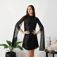 Modelo luciendo un vestido negro brillante con mangas largas y sueltas, posando en un ambiente moderno.