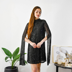 Modelo luciendo un vestido negro brillante con mangas largas, en un entorno moderno con plantas y decoración minimalista.