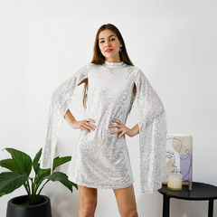 Modelo luciendo un vestido corto plateado con mangas largas y un diseño brillante, posando frente a una planta y una obra de arte.